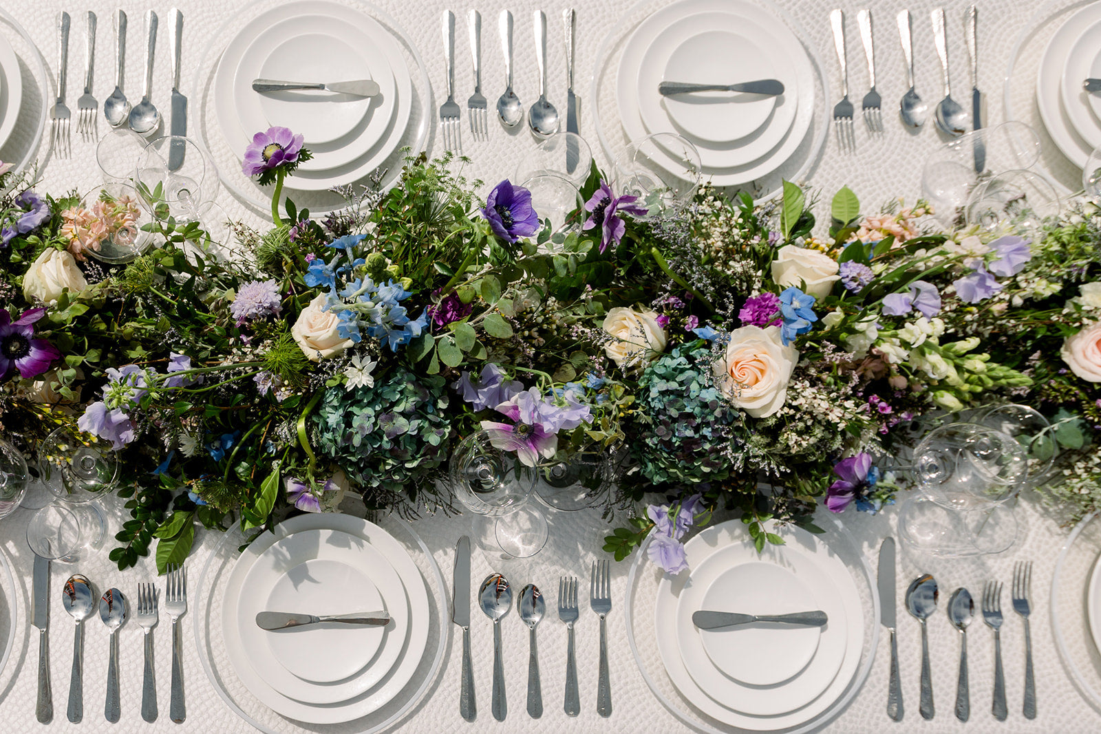 Elegant table setting with floral centerpieces, white plates from borrowed BLU's Reactive Dinnerware Collection, and forged silverware on a white tablecloth.